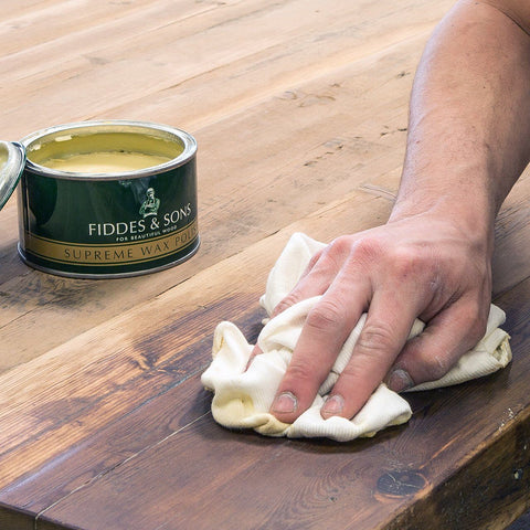 A hand rubbing a cloth over wood with an open tin of wax for wood